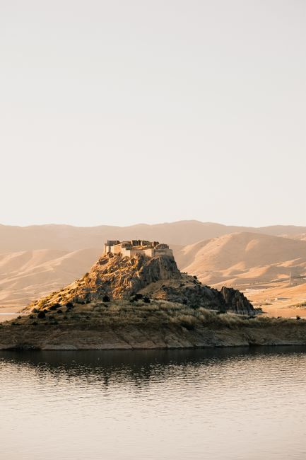 Ancient fortress atop a rocky hill surrounded by serene water and mountains.