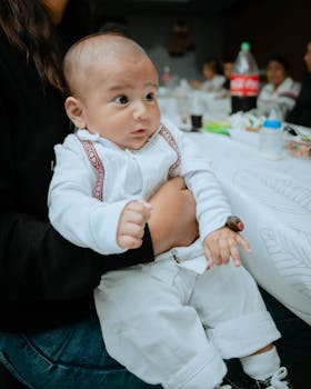 A close-up of a baby being held at a family gathering indoors, with people and food in the background.