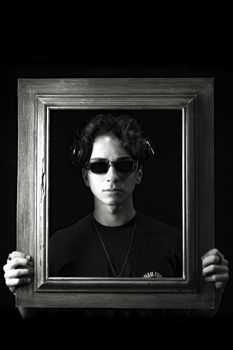 Black and white portrait of a person with sunglasses holding a wooden frame, headphones on.