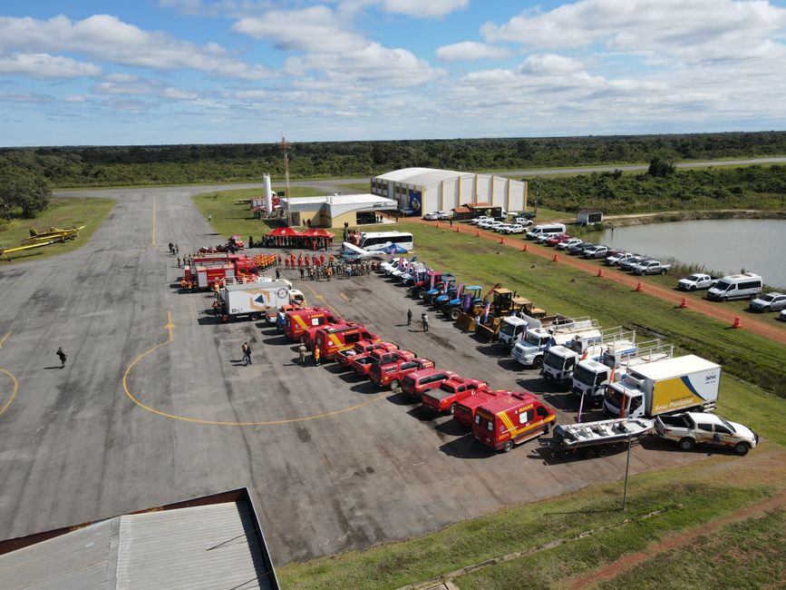 Aerial view of firefighting equipment and vehicles lined up at an airfield. Clear sky, organized deployment.