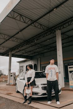 A stylish young couple poses with a car at a vintage gas station, showcasing modern fashion in a rustic setting.
