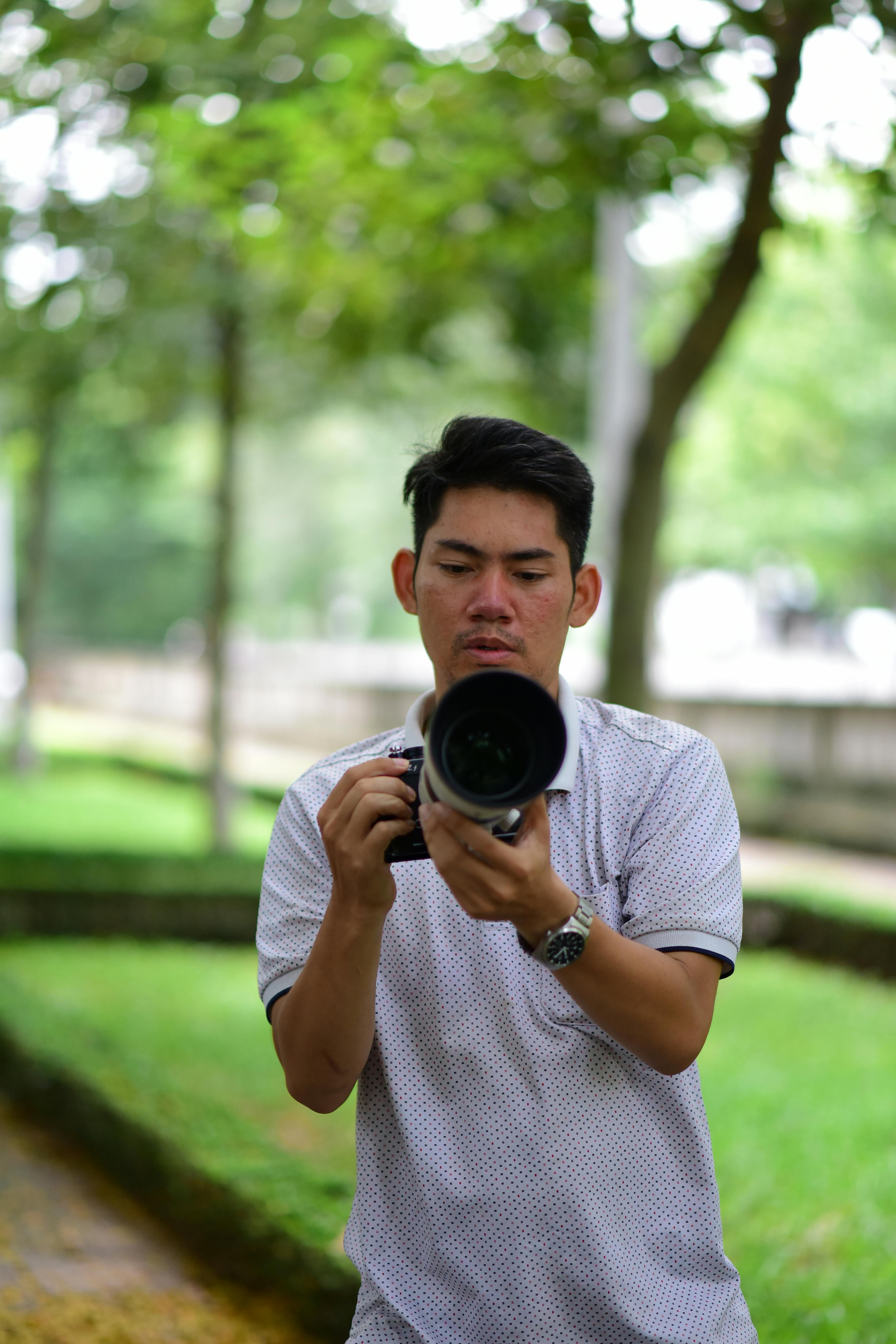 A photographer in a casual shirt adjusting camera settings outdoors with greenery in the background.