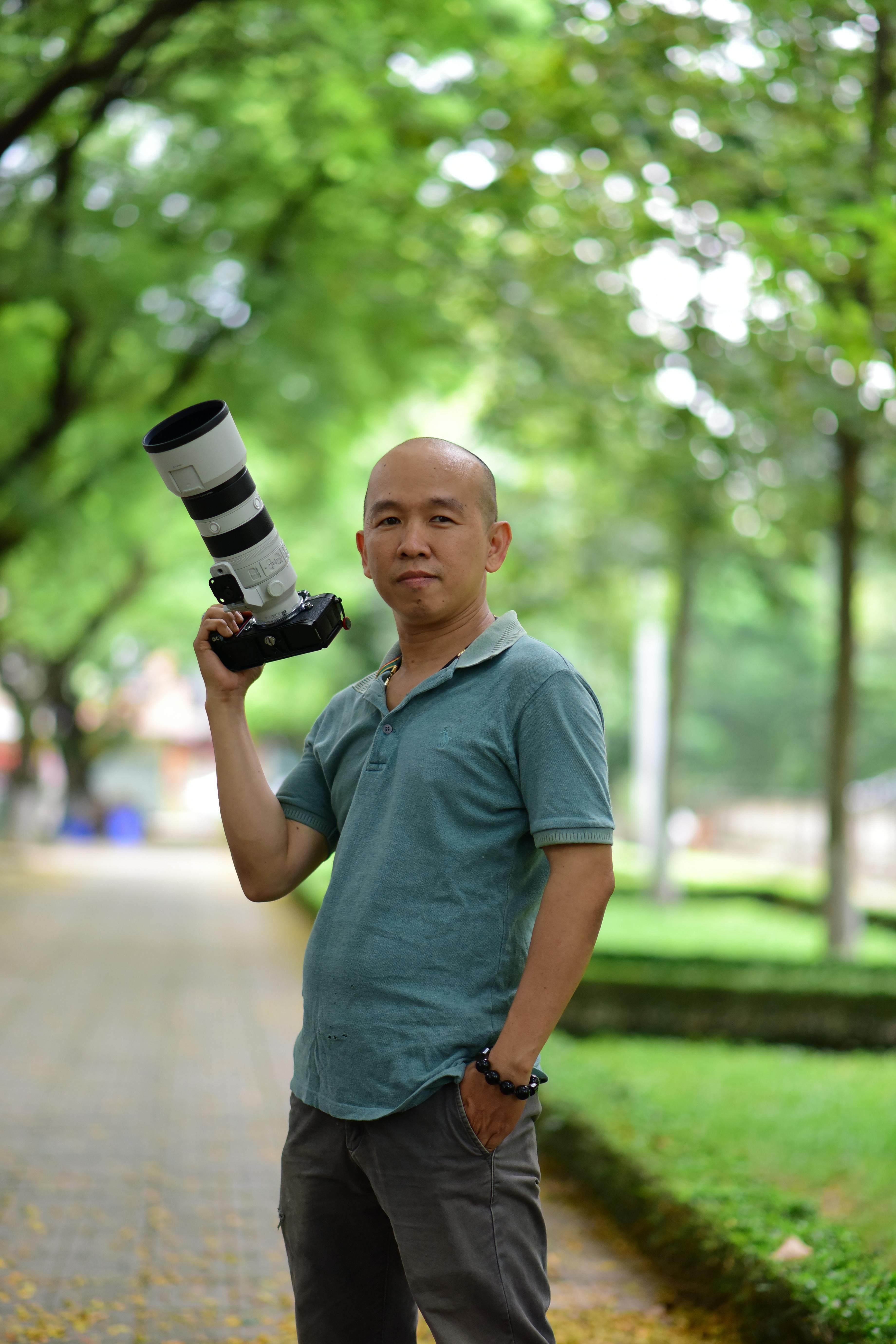 Portrait of a photographer holding a telephoto lens outdoors in a lush, green park setting.