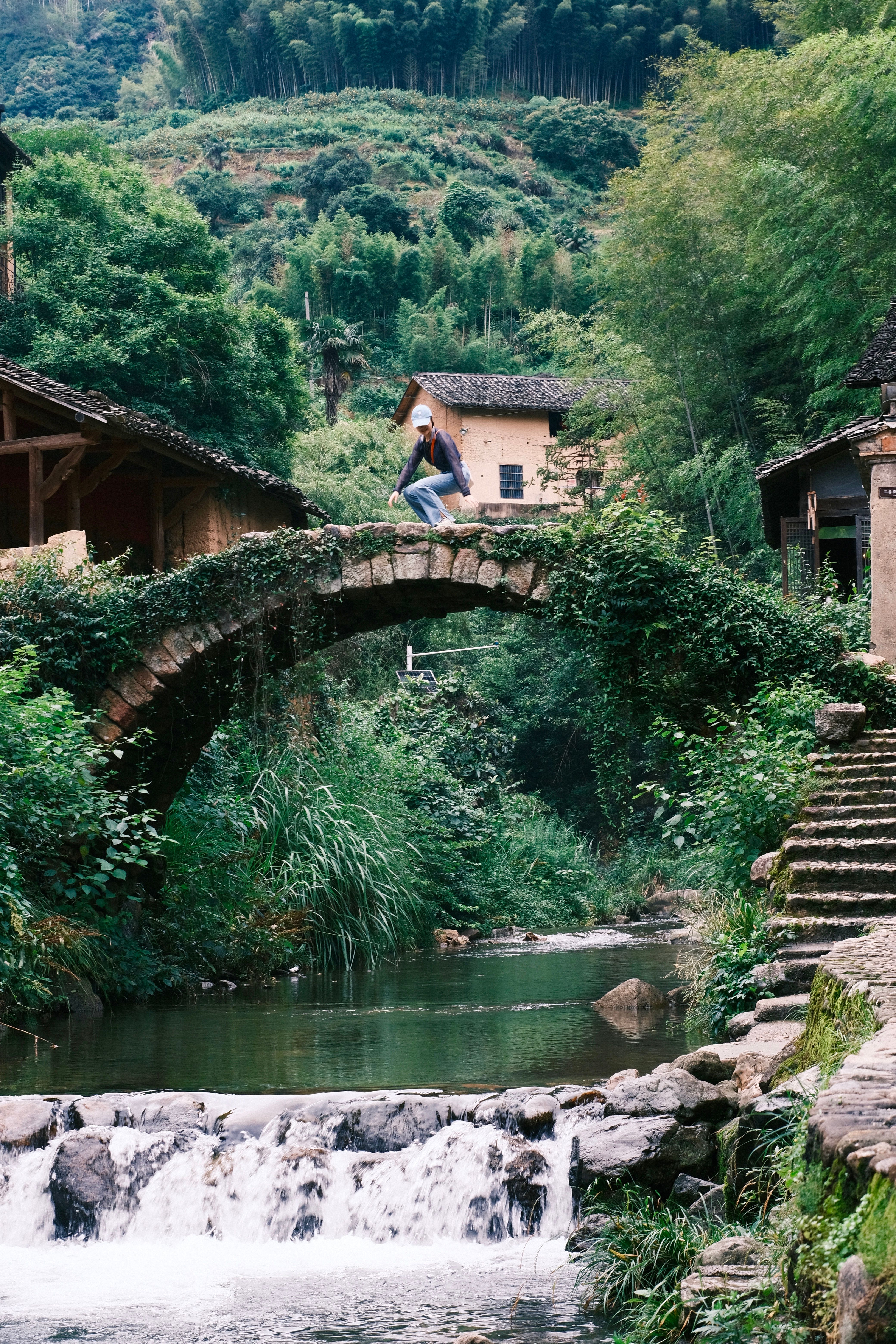 Serene view of an Asian village with a rustic stone bridge over a calm stream surrounded by lush greenery.