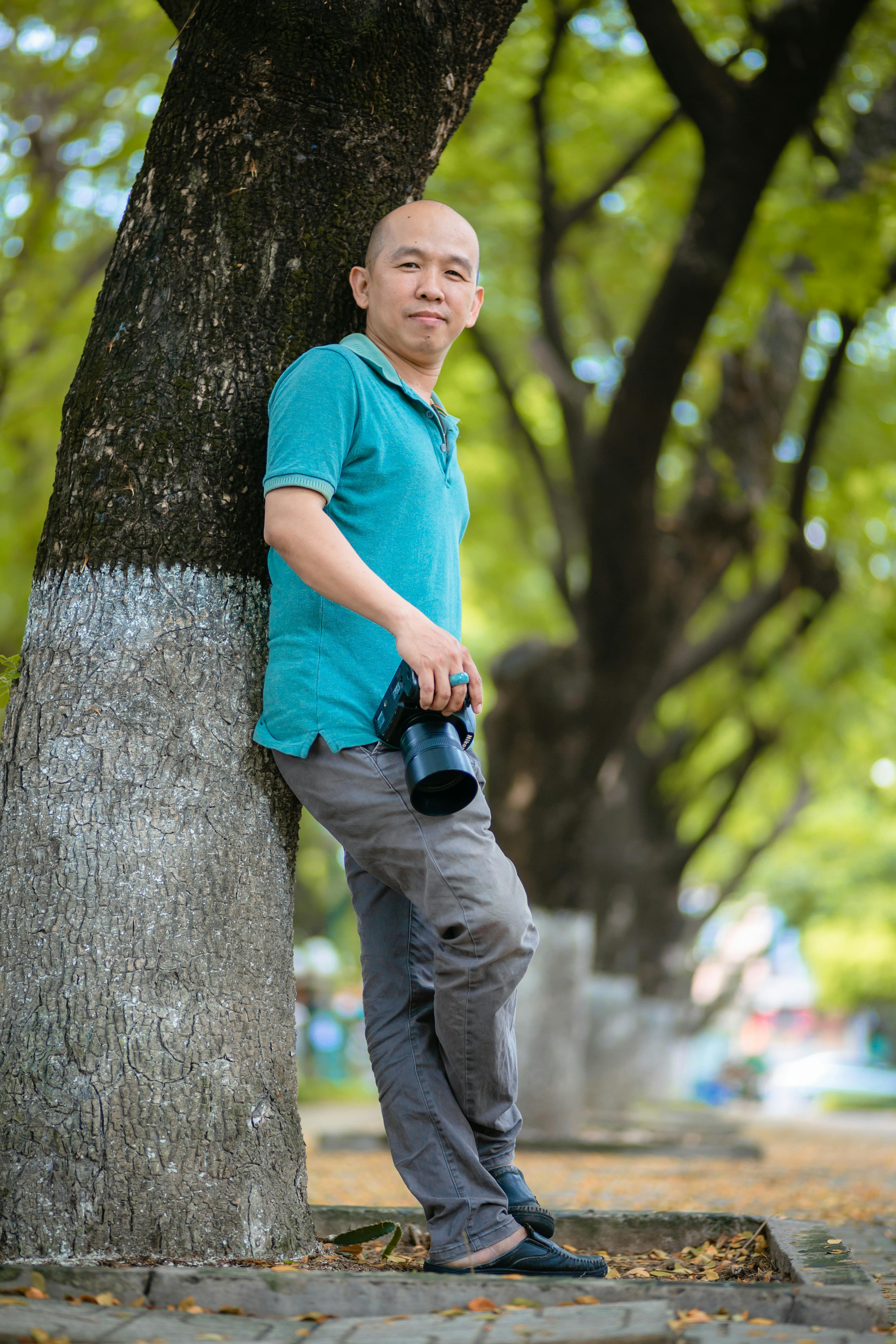 Безкоштовне Дорослий чоловік спирається на дерево та тримає камеру у парку просто неба. стокове фото