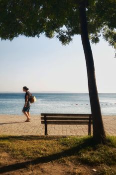 Person walking alone by the ocean on a quiet sunny day with a bench in view.