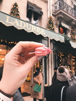 A hand holding a pink macaron with Ladurée storefront in the background.
