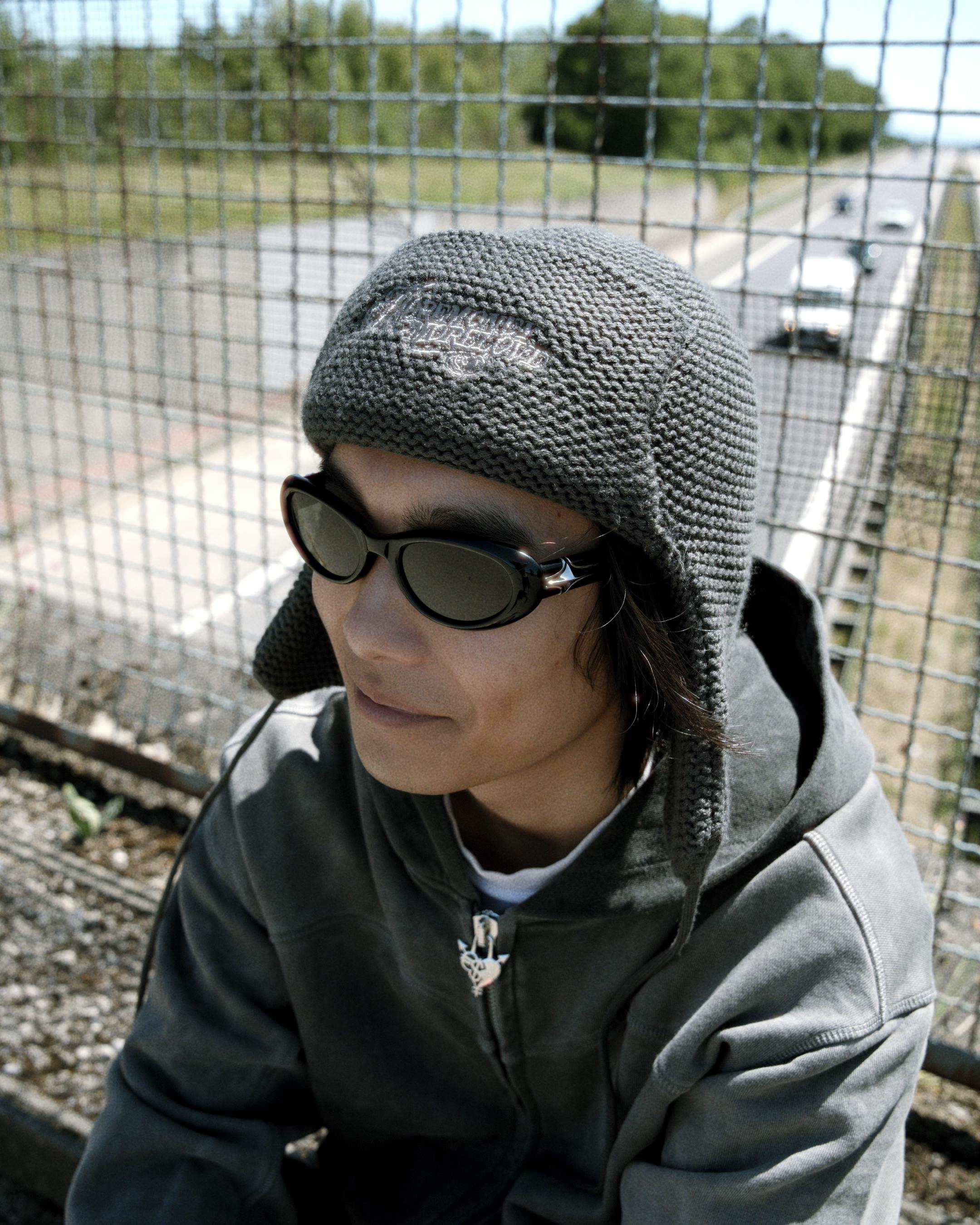 Gratis Seseorang mengenakan kacamata hitam dan topi rajut duduk di dekat pagar pinggir jalan pada hari yang cerah. Foto Stok