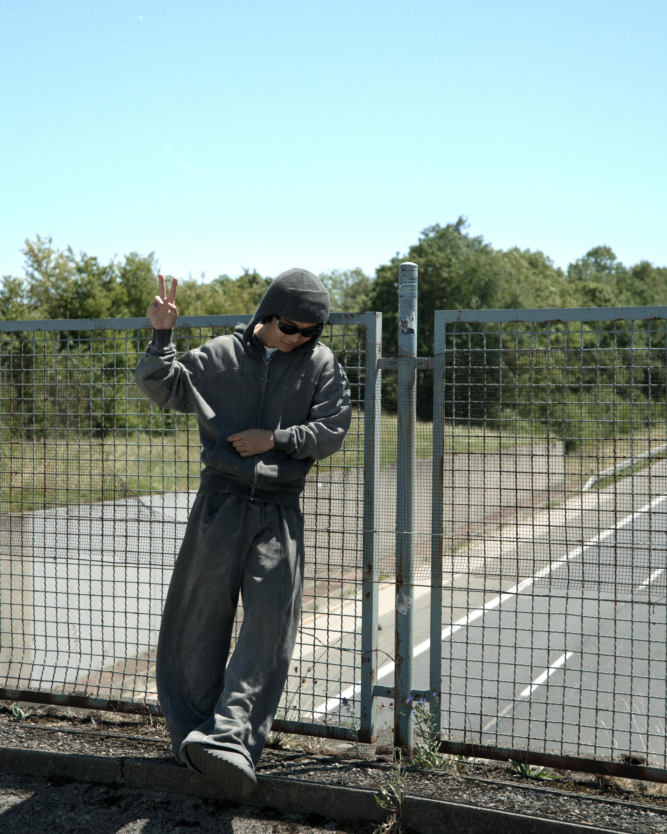 Free Person in casual wear poses near a fence by a road, enjoying a sunny day. Stock Photo