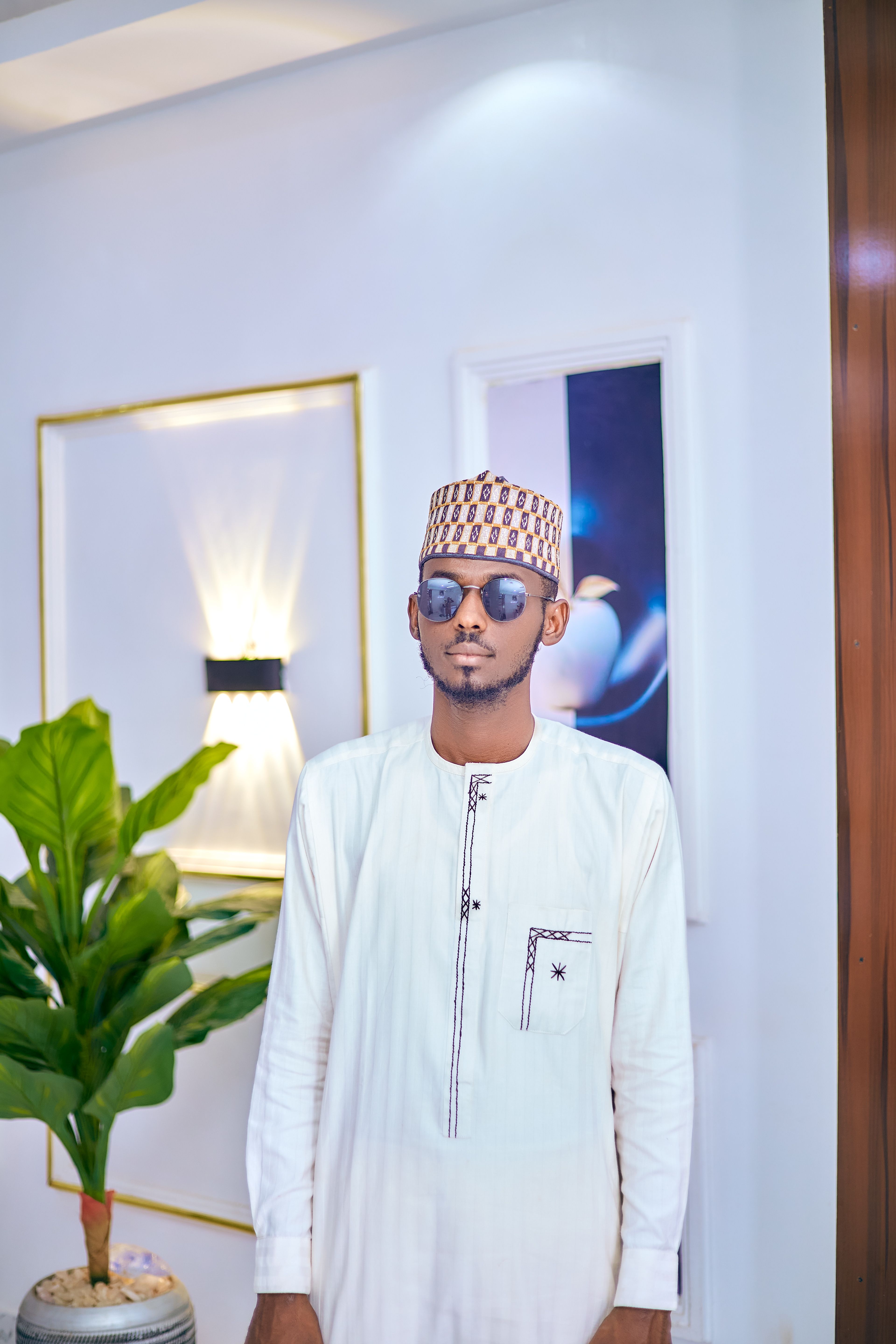 Man in white traditional attire with sunglasses indoors, surrounded by decor.