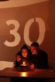 A couple celebrates a cozy 30th birthday with a candlelit cake indoors.