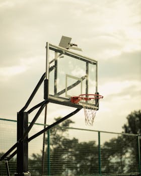 A glass basketball hoop stands outdoors against a cloudy sky, creating a moody sports scene.