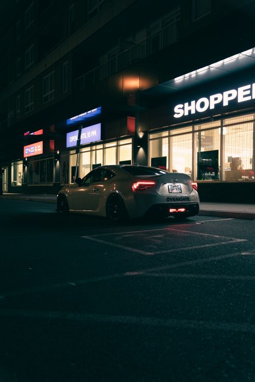 Free A stylish car parked under city lights at night, in front of a modern shopping center. Stock Photo