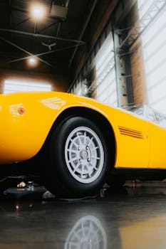 Low angle view of a vintage yellow sports car parked indoors with natural light streaming through large windows.