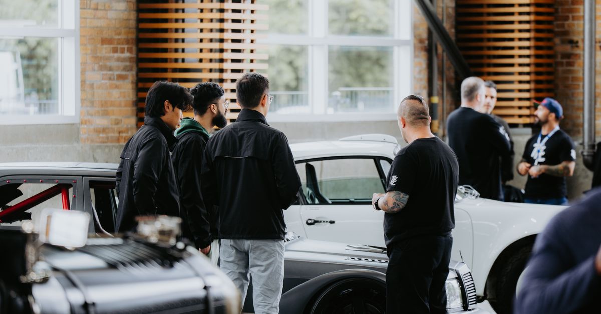Men gather around vintage cars in an indoor setting, discussing automotive designs.