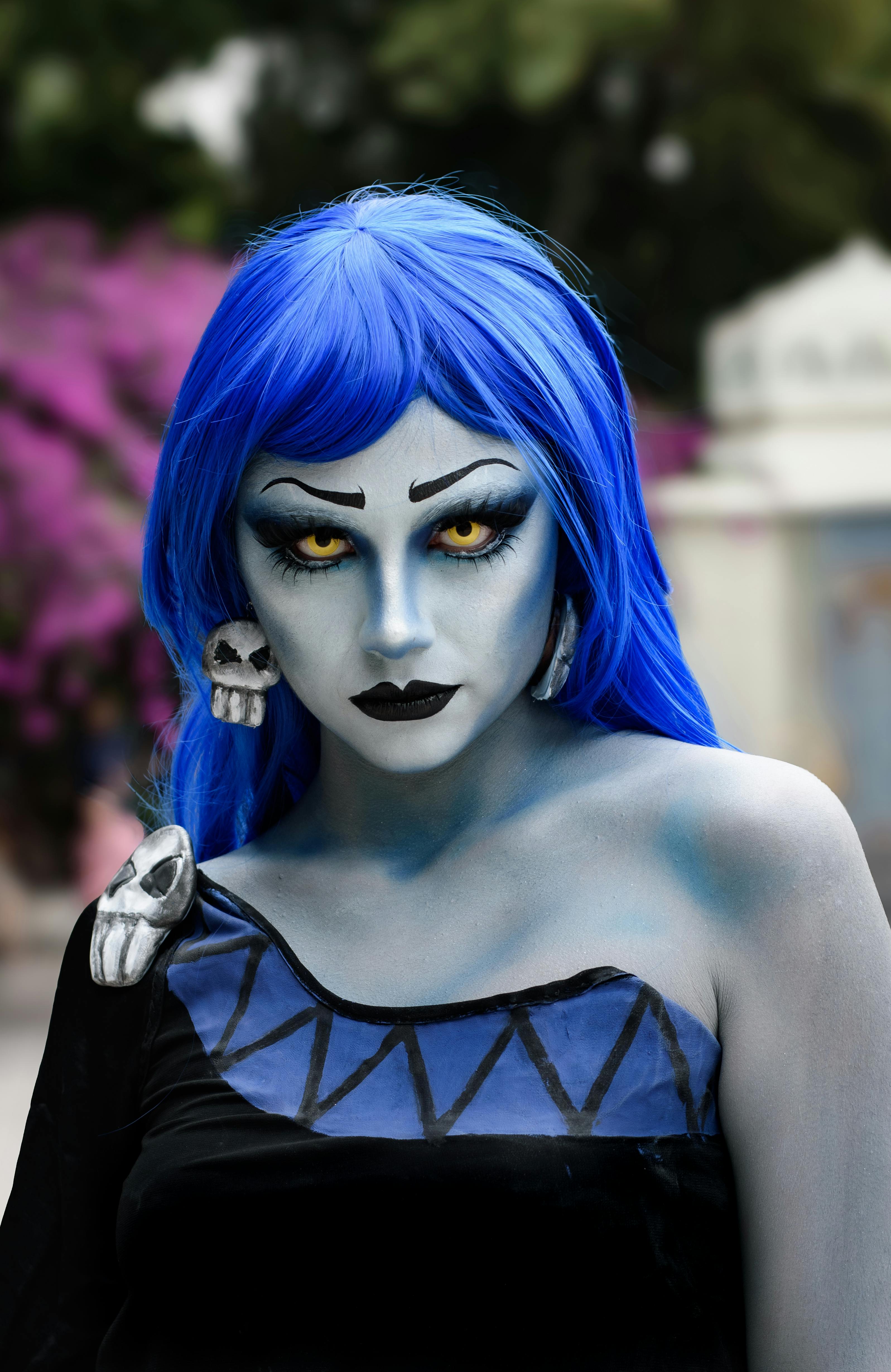 A striking cosplay portrait featuring a person with blue hair and intense makeup, set against a blurred outdoor backdrop.