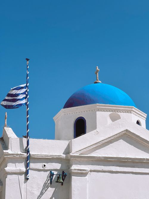 Free Iconic Greek church with a blue dome and national flag under a clear blue sky. Stock Photo