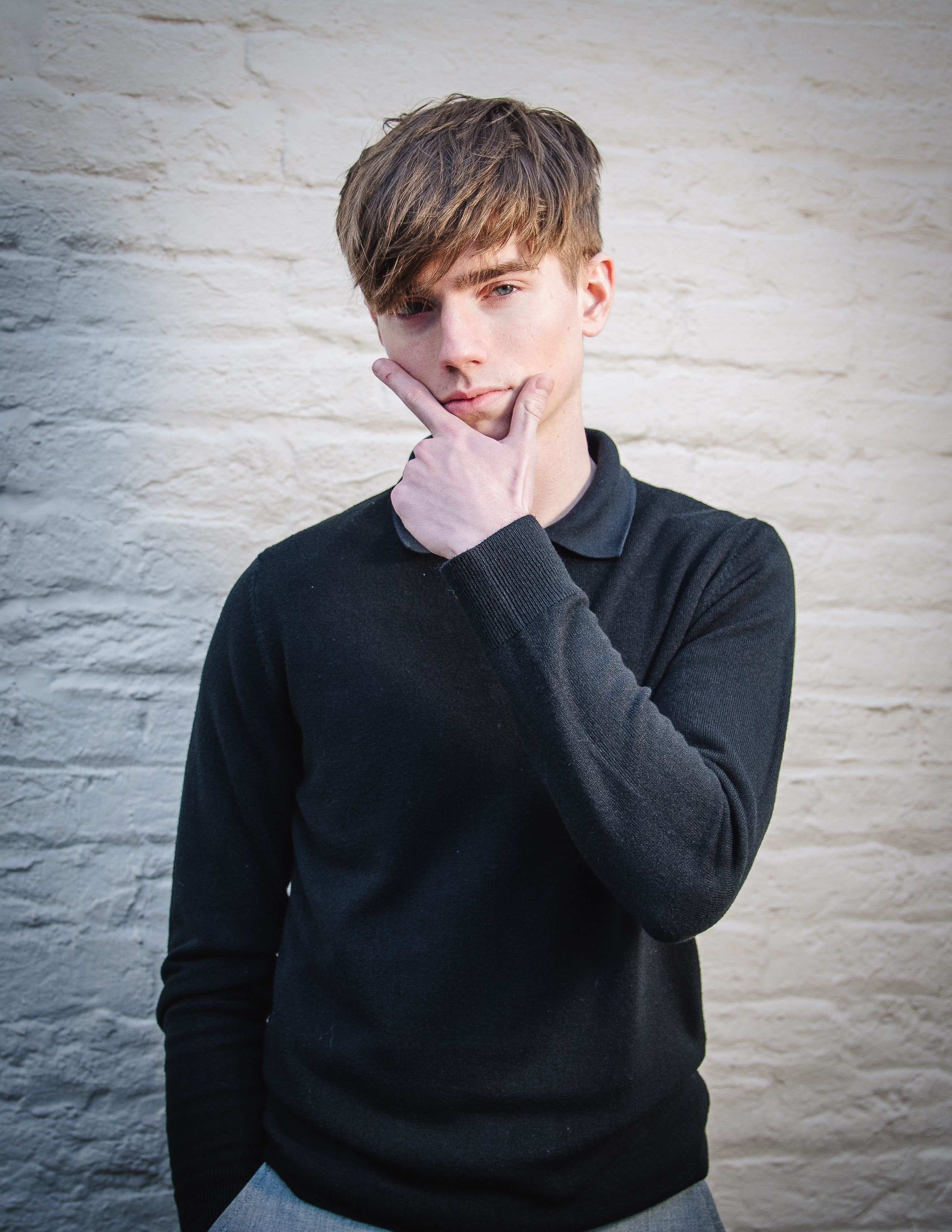Young man in casual attire striking a thoughtful pose against a textured brick wall background.