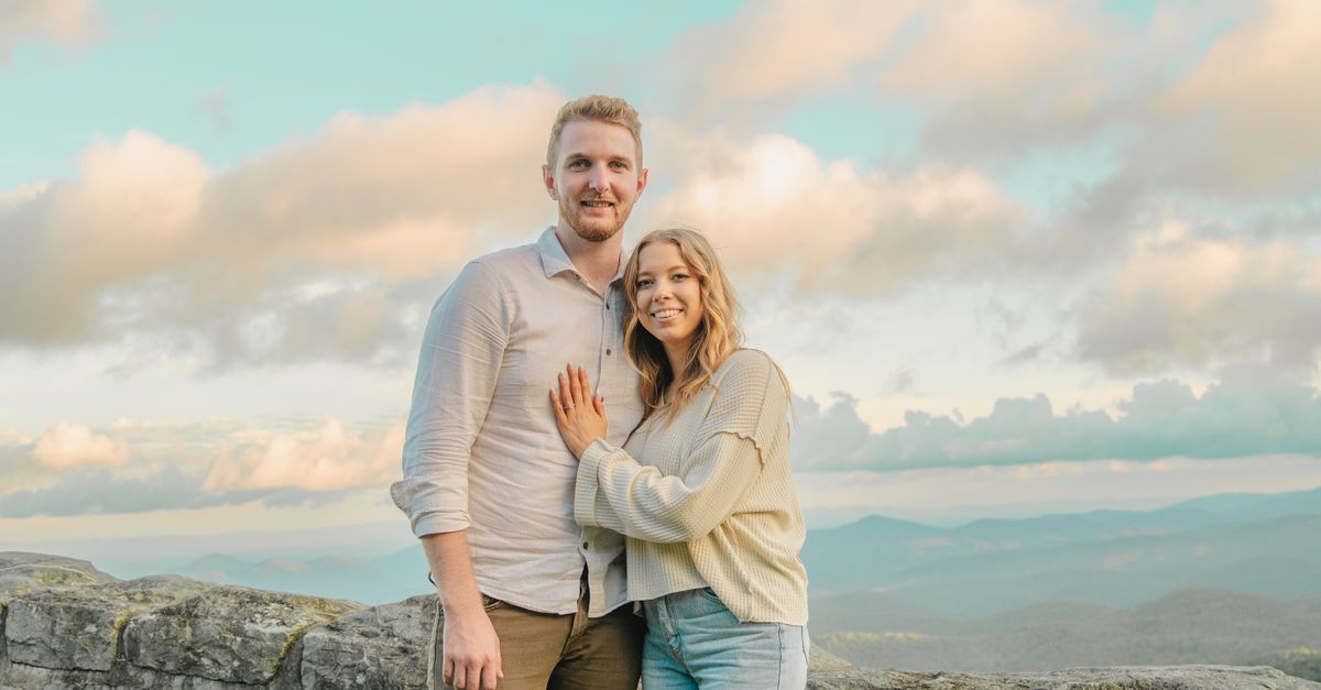 A smiling couple embraces with a stunning mountain landscape in the background, captured at sunset.