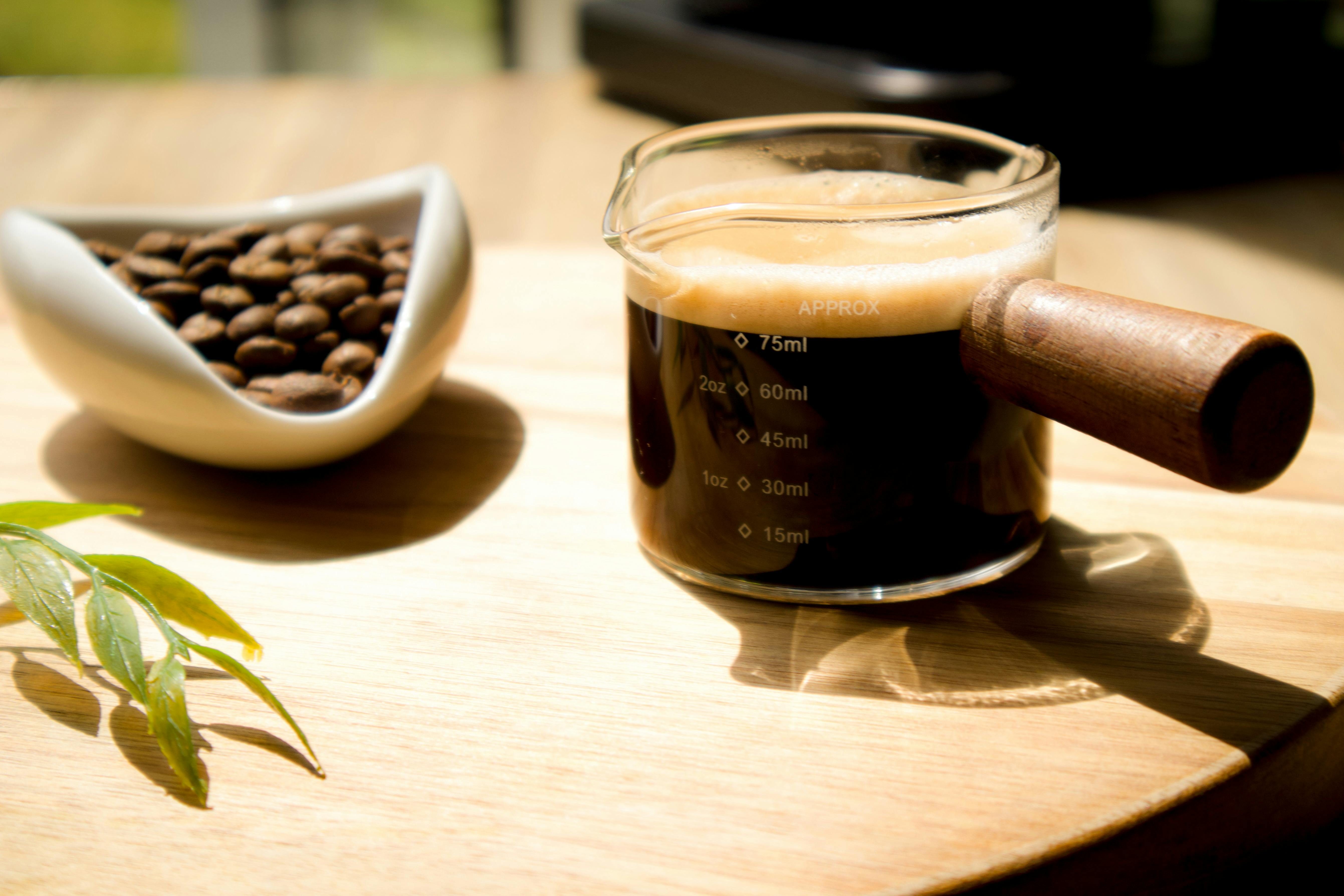 Glass espresso cup with crema beside a bowl of coffee beans on a sunny wooden table.