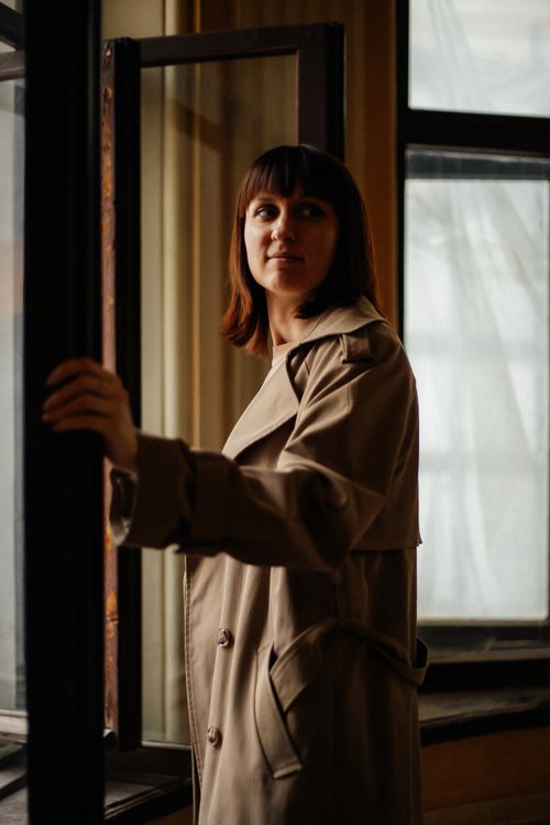 Free Thoughtful woman in a beige trench coat standing by an open window indoors. Stock Photo