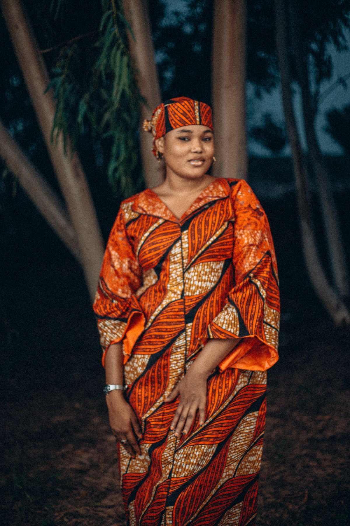 Woman in an orange leaf-print Ankara outfit among trees at dusk