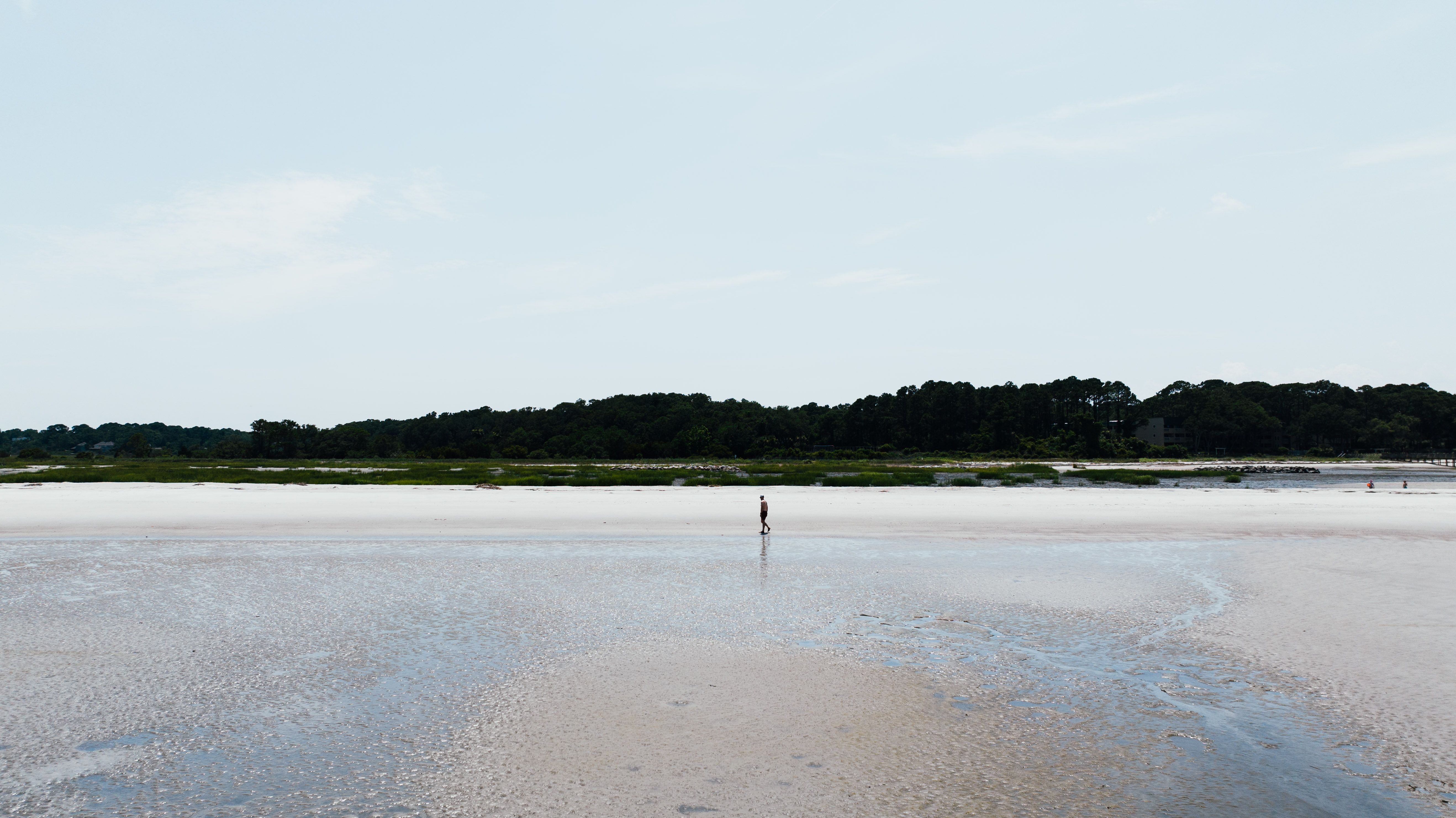 A lone person stands on a wide sandy beach at Hilton Head Island under a clear sky.