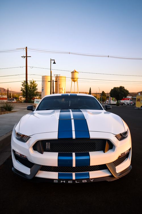 Free White Shelby sports car with blue stripes at sunset, parked on street with industrial background. Stock Photo
