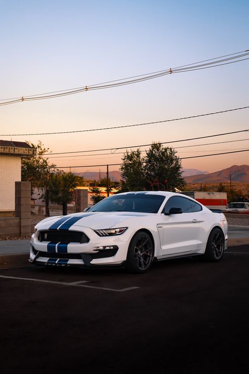 Free Sleek white sports car with blue racing stripes parked in a city lot at sunset. Stock Photo