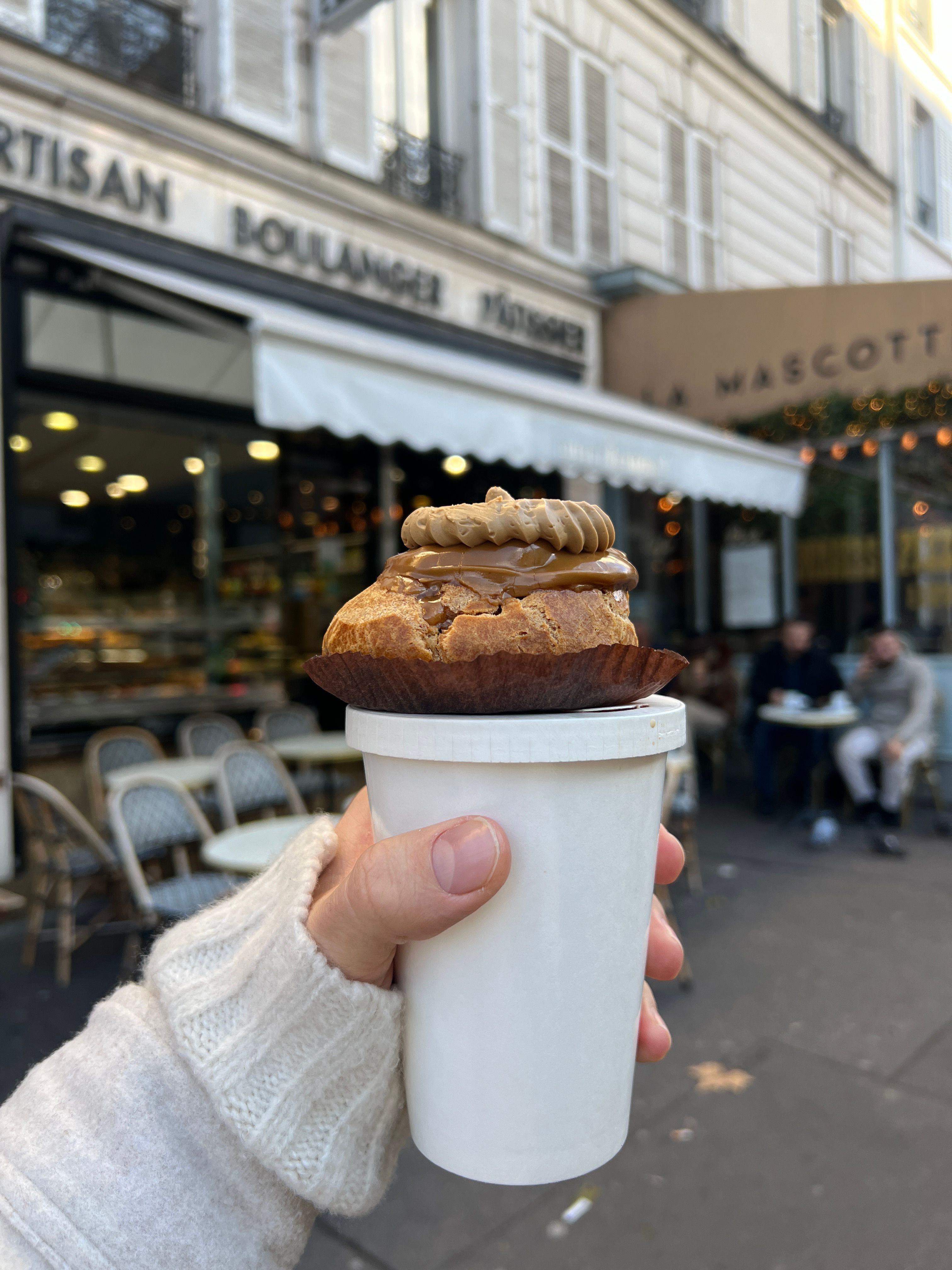 Hand holding coffee and pastry outside Parisian bakery, cozy street view.