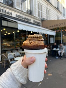 Hand holding coffee and pastry outside Parisian bakery, cozy street view.