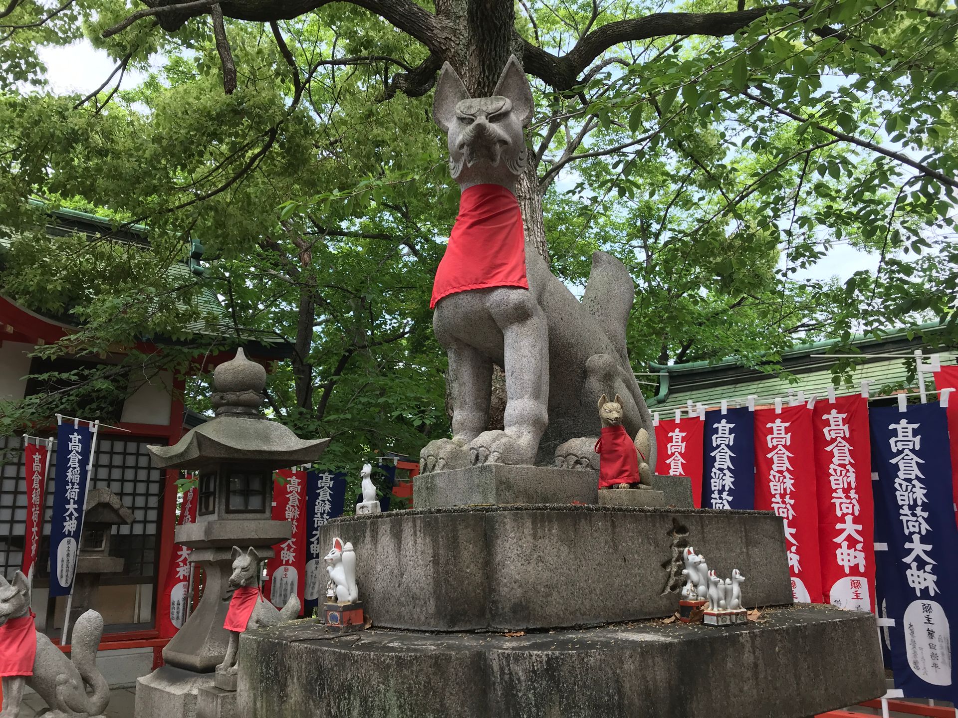 A stone fox statue wears a red bib at a Japanese Inari shrine surrounded by trees and banners.