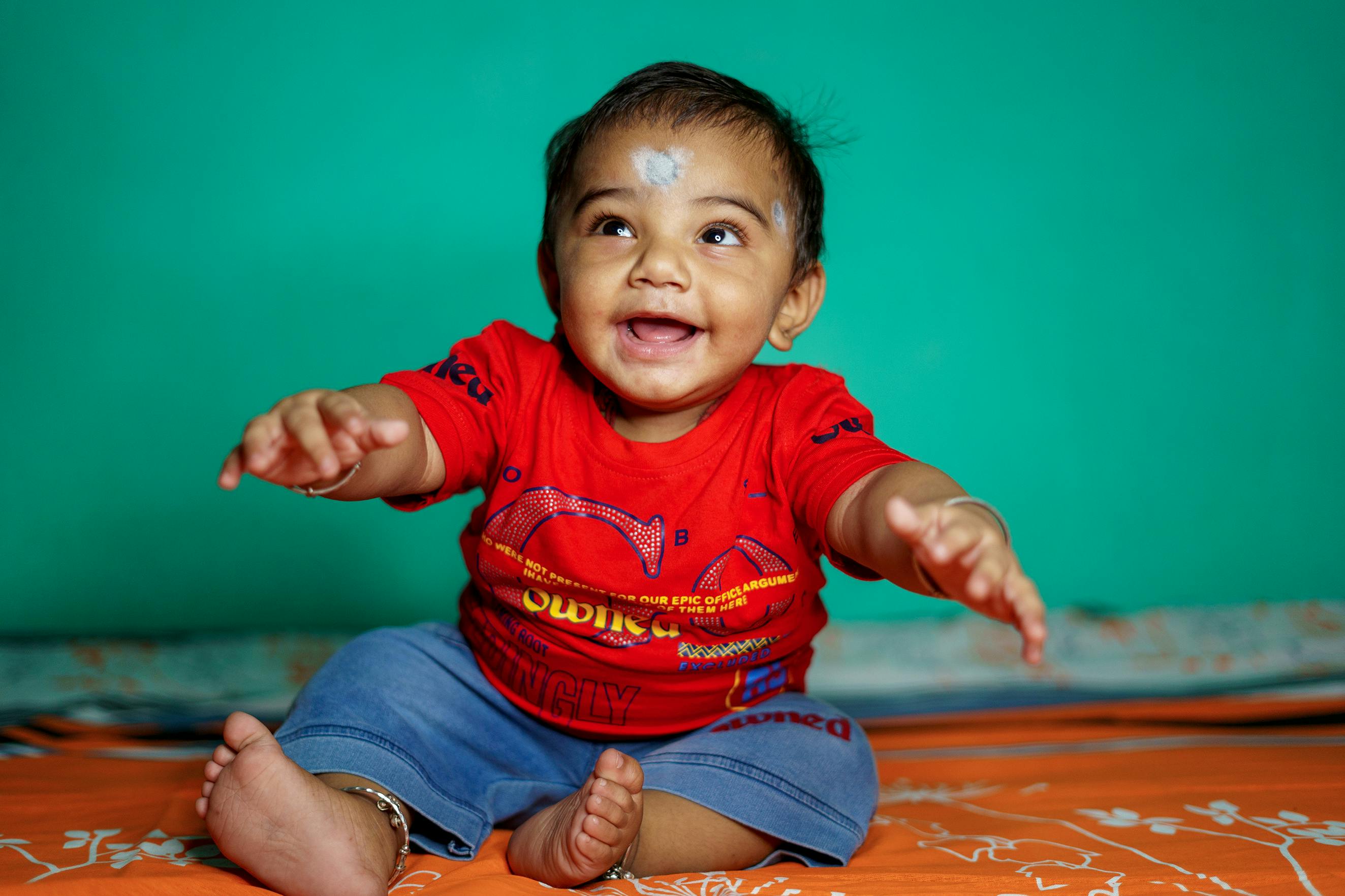 Adorable baby boy sitting indoors, reaching out with a joyful smile.