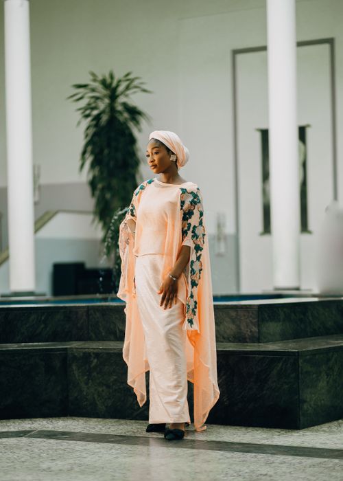 Free Sophisticated woman in peach dress with shawl standing indoors, exuding grace and style. Stock Photo