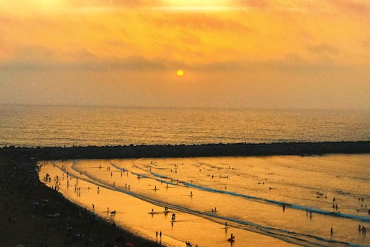 A stunning sunset over a crowded beach with swimmers and surfers enjoying the waves.