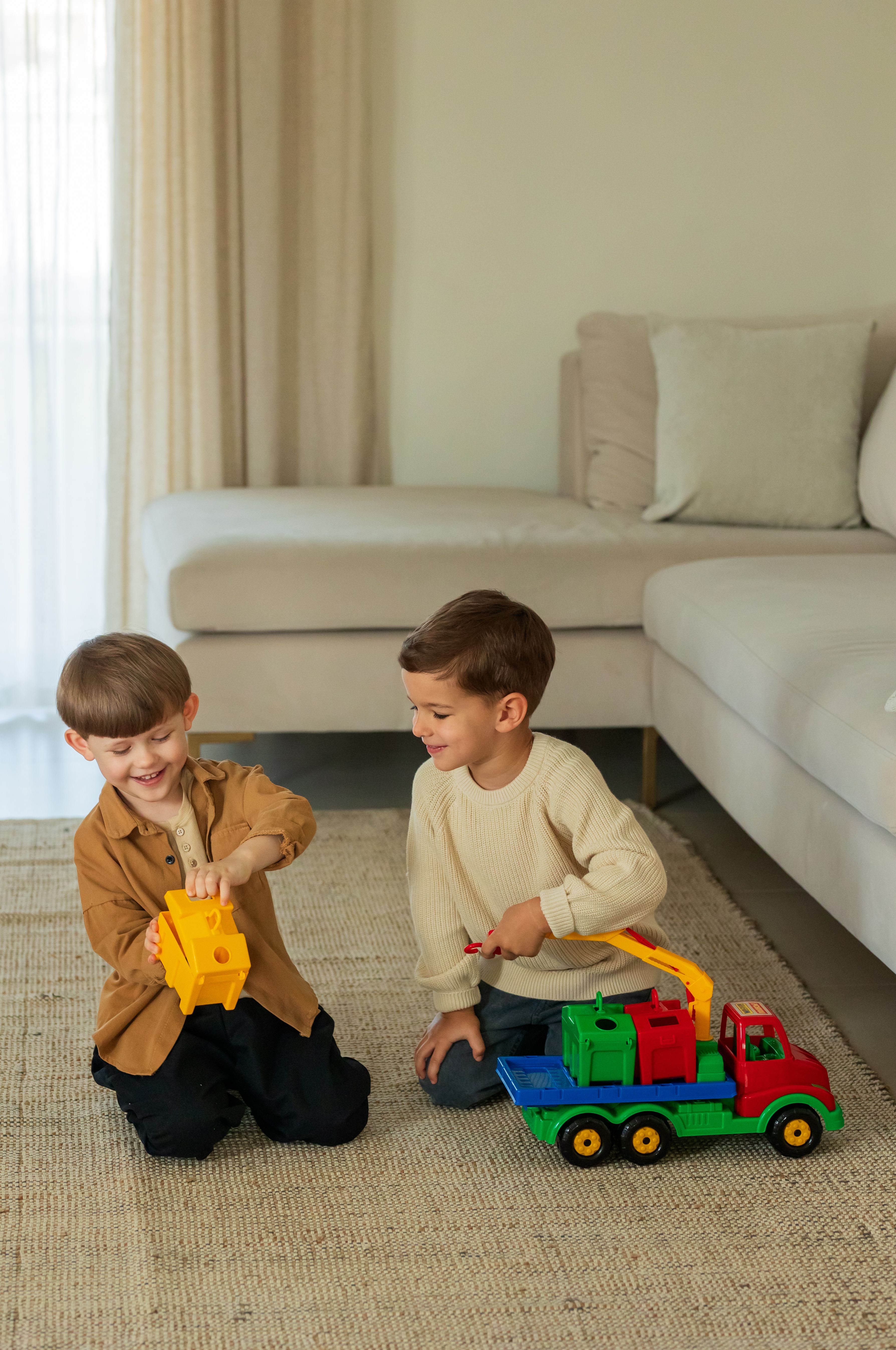 Two boys enjoy playing with colorful toy trucks on a carpeted floor in a cozy living room setting.