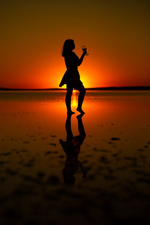 Free A woman holding a glass, silhouetted against a vibrant sunset reflected on water, creating a serene atmosphere. Stock Photo
