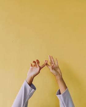 Two hands gracefully reach up against a bright yellow wall, creating a minimalist and artistic composition.