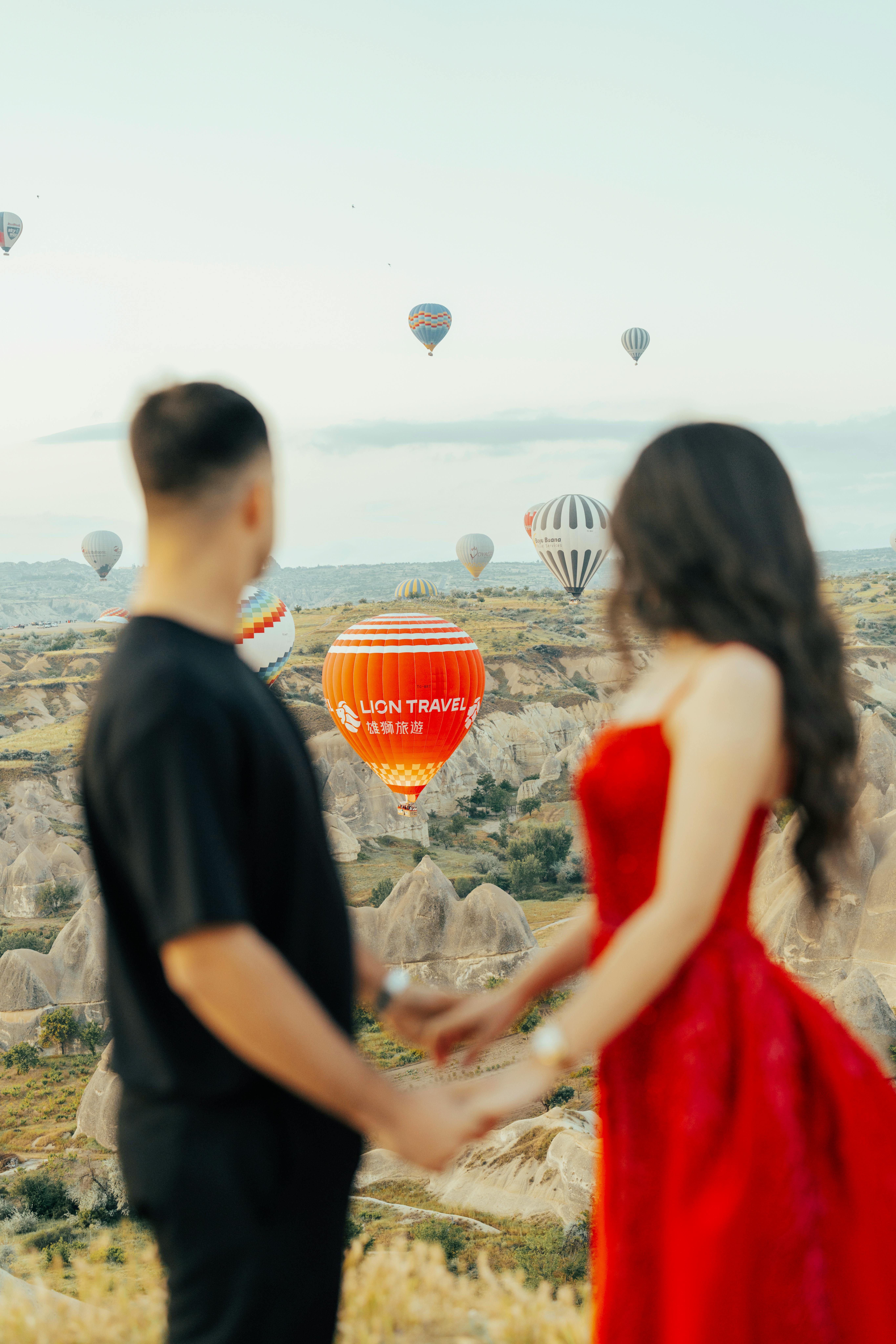 Couple holding hands and admiring hot air balloons over Cappadocia's scenic landscape in Türkiye.
