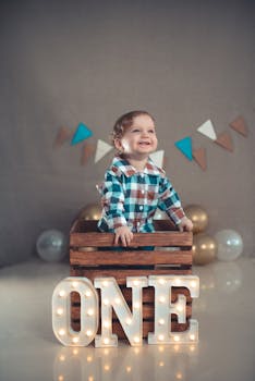 Smiling toddler in plaid shirt poses for first birthday photo with lit 'ONE' sign and balloon decor indoors.