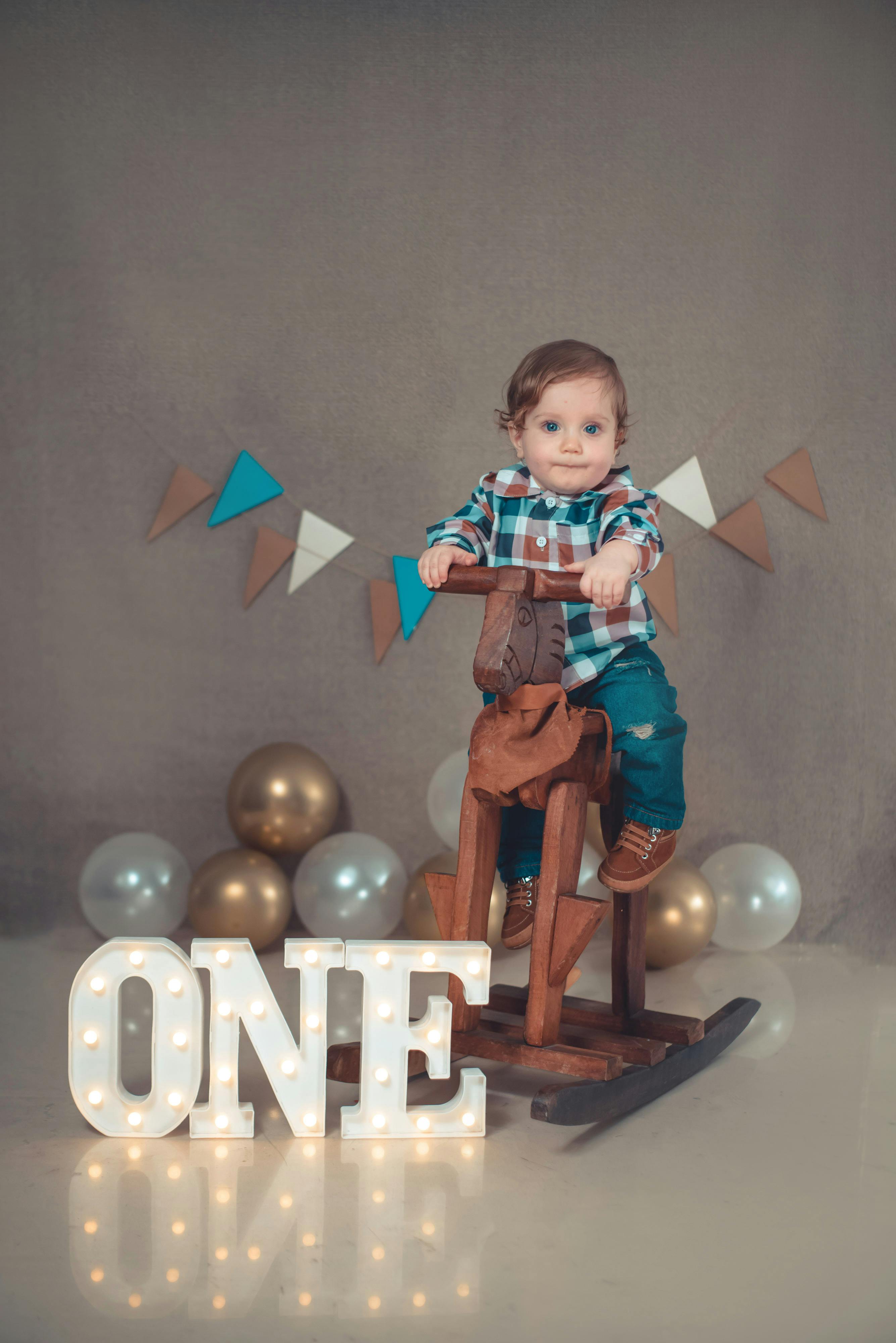 Adorable baby boy on a rocking horse celebrating his first birthday indoors, surrounded by balloons and decor.