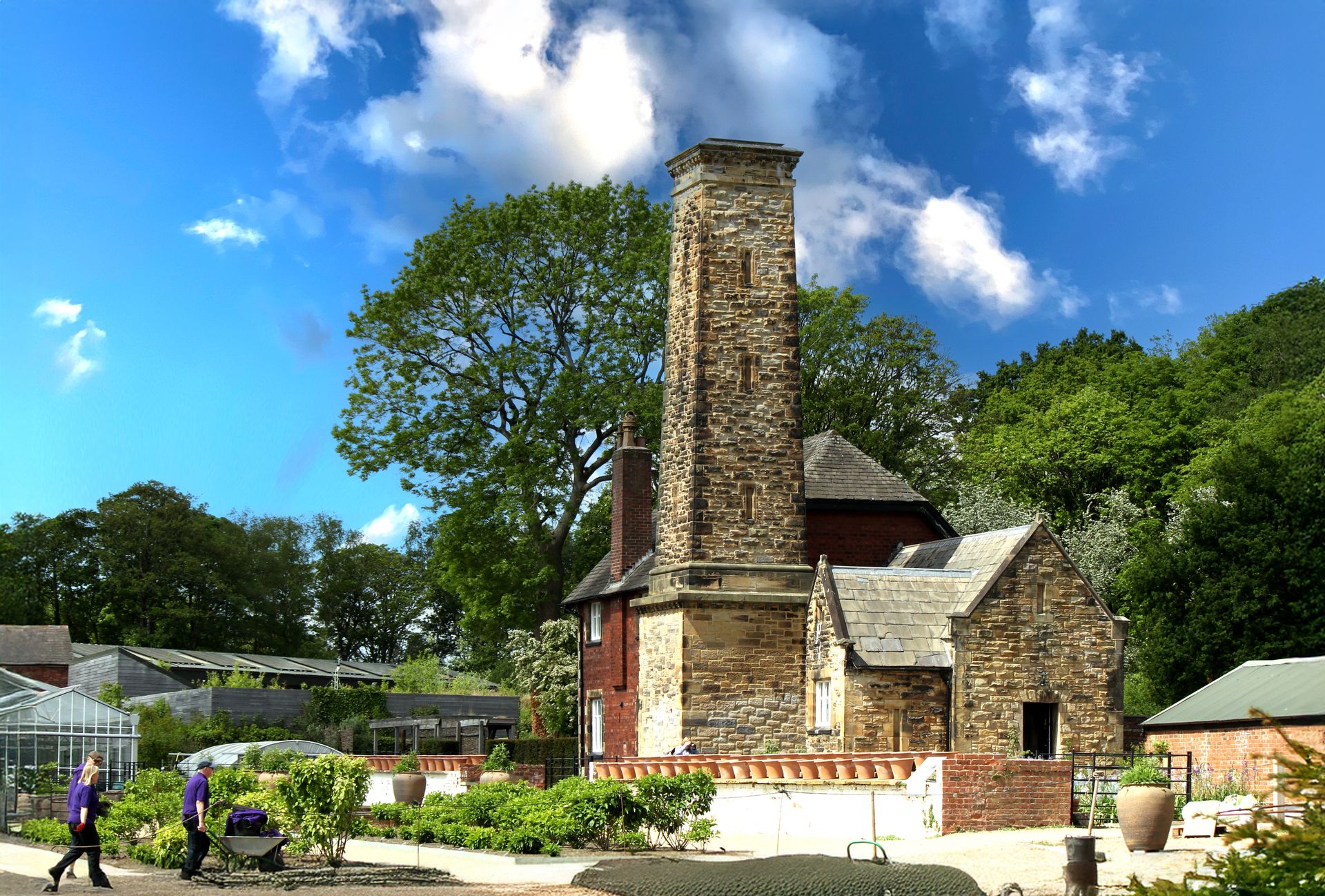 Idyllic view of a historic stone tower surrounded by green trees and gardeners in an English countryside garden.