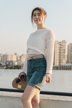 Stylish woman in casual outfit by urban waterfront, sunlit city backdrop.