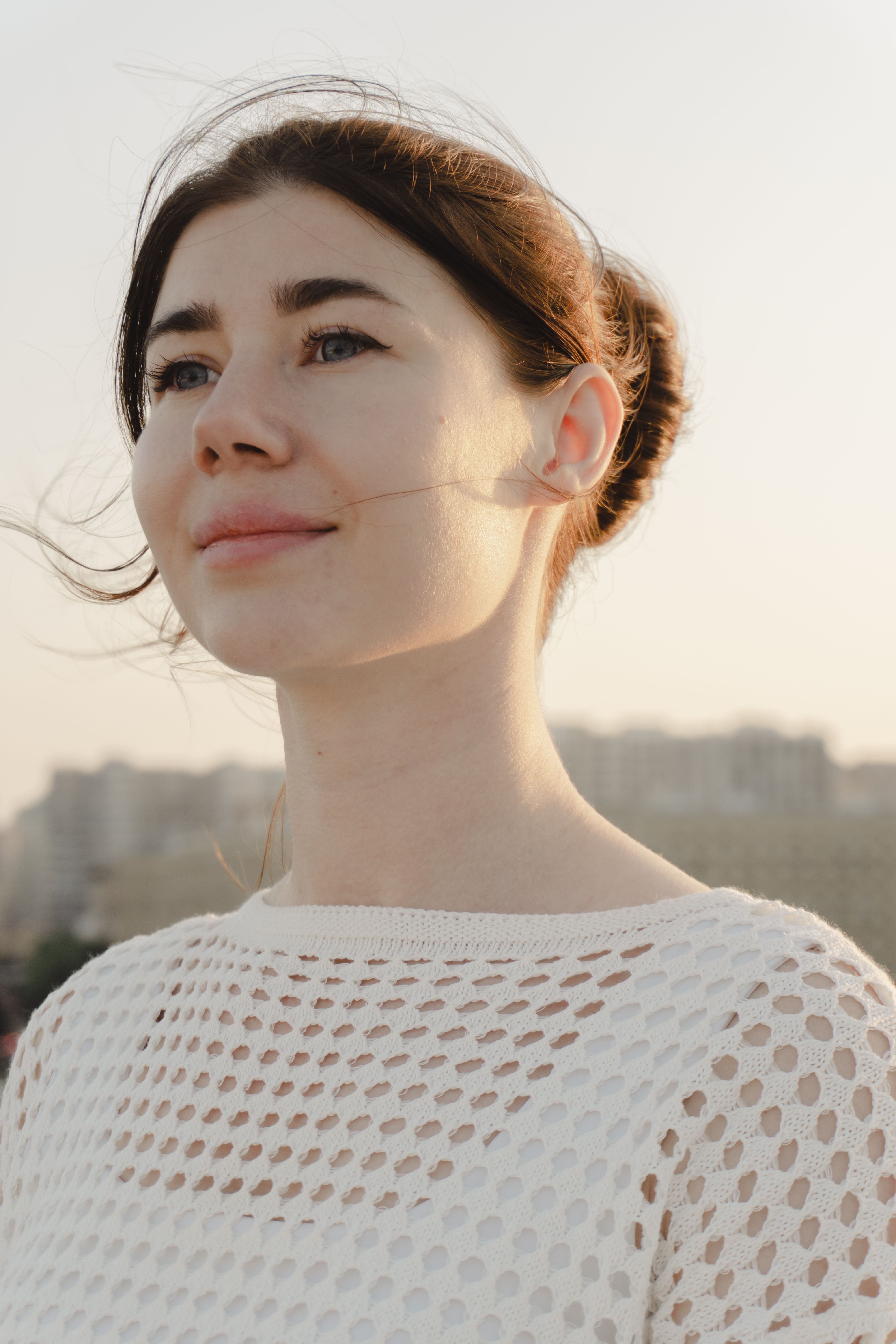 A woman with a thoughtful expression in a white mesh top outdoors against a blurred background.