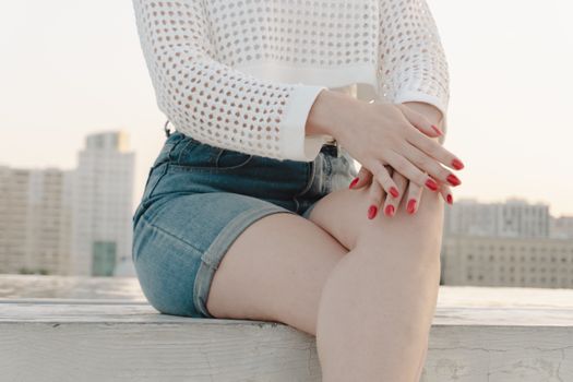 Close-up of a woman in denim shorts and knitted sweater on a rooftop with cityscape.