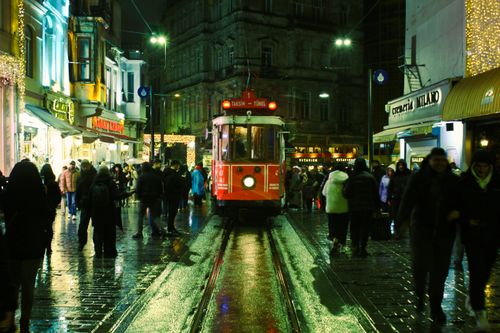 Free Bustling night scene on Istiklal Avenue featuring iconic red tram and streetlights reflecting on wet pavement. Stock Photo