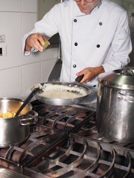 Professional chef seasoning a creamy sauce in a stainless steel pan on a commercial stove.
