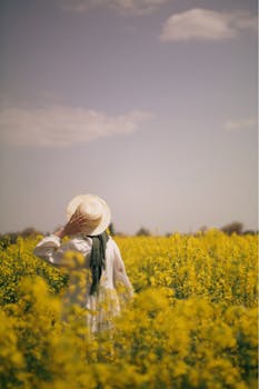 A woman in a straw hat walking through a vibrant yellow flower field on a sunny day.