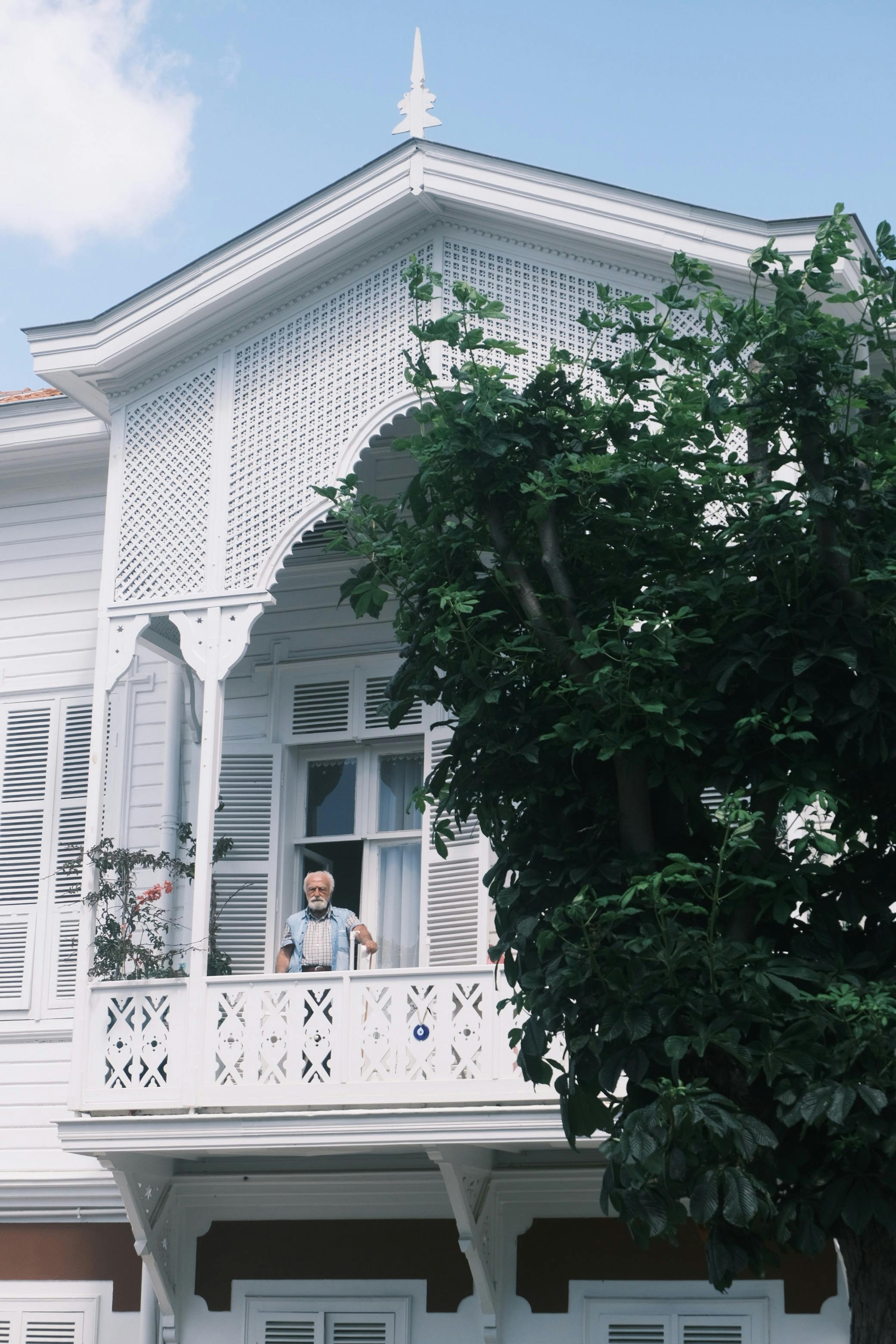 A senior man stands on a historic balcony with intricate white architecture and lush greenery.