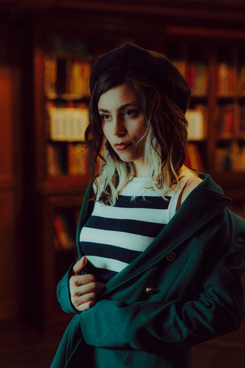 Free Artistic portrait of a woman in a beret, wearing classic stripes, standing in a dimly lit library. Stock Photo